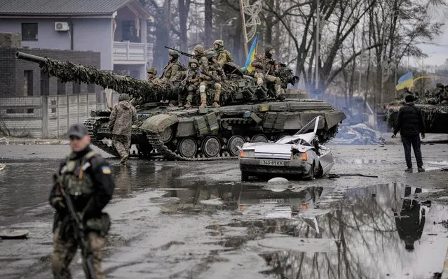 A man stands next to a civilian vehicle that was destroyed during fighting between Ukrainian and Russian forces that still contains the body of the driver as Ukrainian servicemen ride past on a tank outside Kyiv, Ukraine, Saturday, April 2, 2022. (Photo by Vadim Ghirda/AP Photo)