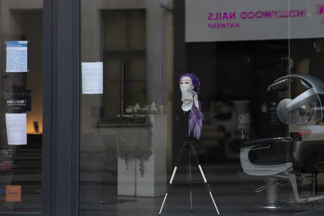 A mannequin head with a scarf around the nose and mouth is placed on a tripod in the window of a hairdressing salon in Antwerp, Belgium, Monday, March 23, 2020. After Belgian Prime Minister Sophie Wilmes announced that hairdressers would still be able to operate, while many other stores were required to be closed during the coronavirus epidemic, some thought it was a bad Belgian joke. Fearing for their health, some hairdressers now are calling on the government to order the closure of all salons without delay. (Photo by Virginia Mayo/AP Photo)