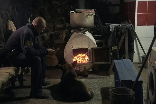 An emergency worker and his dog warm up in front of a wood-burning oven in a shelter in Bakhmut, the site of the heaviest battles with the Russian troops, in the Donetsk region, Ukraine, Friday, December 9, 2022. (Photo by LIBKOS/AP Photo)