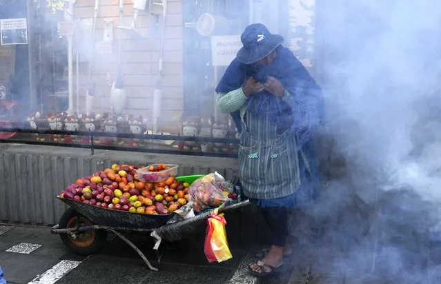A fruit vendor covers her nose and mouth amid tear gas fired by police during clashes with protesting educators near the government palace in La Paz, Bolivia, Wednesday, April 12, 2023. Teachers are demanding higher pay and protesting the new curriculum at public schools. (Photo by Juan Karita/AP Photo)