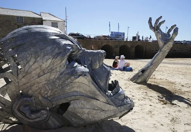 A metal sculpture by artist Ruscoe, which depicts a sinking man, sits amidst sunbathers on a beach outside the G7 meeting in St. Ives, Cornwall, England, Sunday, June 13, 2021. (Photo by Jon Super/AP Photo)