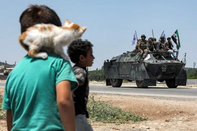 A boy with a kitten perched on his shoulder looks on as Turkish-backed Syrian rebel fighters from Tal Rifaat ride in, and above, an armoured personnel carrier (APC) moving in a military column headed for the Azaz frontlines on the road from Afrin, in Qatmah in the countryside of Syria's northern Aleppo province on July 2, 2022. (Photo by Bakr Alkasem/AFP Photo)