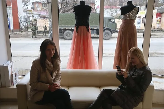 Two young women sit in a shop that sells evening gowns as heavily-armed soldiers displaying no identifying insignia stand outside in a street in the city center on March 1, 2014 in Simferopol, Ukraine. Similarly dressed soldiers stationed themselves around the Crimean Parliament building earlier in the day in what seems to be a growing military occupation of the city, which is the Crimean capital. (Photo by Sean Gallup/Getty Images)