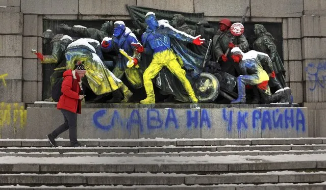 A man walks by a monument of the Soviet army spray painted in yellow and blue, the colors of the Ukrainian flag, and writing reading “Glory of Ukraine”, in Sofia, Monday, February 28, 2022. In a flash mob on Monday, several protesters gathered at the monument and wrote with colored sprays slogans protesting against Russia's invasion of Ukraine. (Photo by Valentina Petrova/AP Photo)