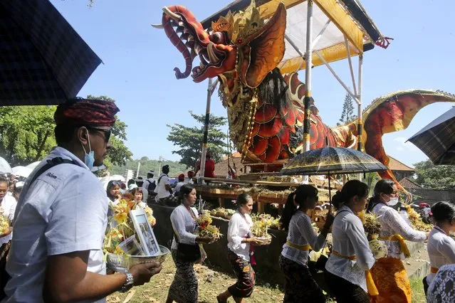 People carry portraits of their dead relatives as they walk past a giant effigy of a mythical animal which will be used in a traditional mass cremation ceremony called “ngaben” on Friday, July 29, 2022, in Padangbai on the island of Bali, Indonesia. Balinese believe that cremating the dead liberates their souls, allowing them to enter the higher world to reincarnate into better beings. (Photo by Firdia Lisnawati/AP Photo)