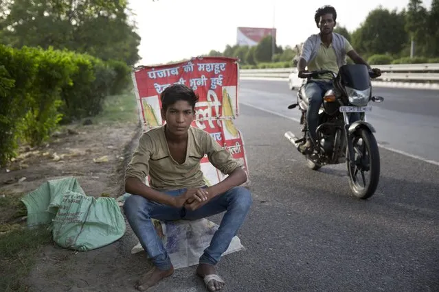 In this Tuesday, June 6, 2017 photo, an Indian boy who sells corn along a busy expressway poses for a photo next to his corn stall as a man on a motorcycle patrols to ensure business runs smoothly in Noida, India. Every 100 meters (330 feet) or so there are children selling corn along this busy expressway on the outskirts of New Delhi. According to India’s 2011 census, boys like him are part of the estimated 8.3 million child laborers In India. Uttar Pradesh state, where Noida is located, alone accounts for 1.8 million of that total. (Photo by Tsering Topgyal/AP Photo)