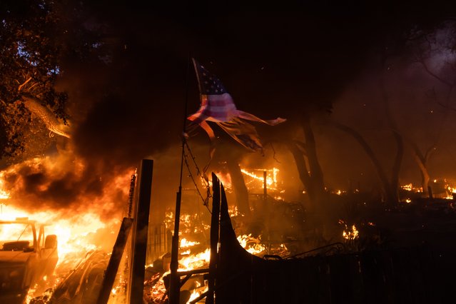 An American flag are seen in front of a house in flames at El Cariso Village in flames as the Airport Fire burns on September 10, 2024 in Lake Elsinore, California. The fast-moving Airport Fire that started on September 9 in Orange County grew up to more than 10,000 acres by this afternoon, fire officials said. (Photo by Apu Gomes/Getty Images)