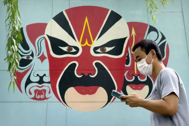A man wearing a face mask to protect against the spread of COVID-19 walks past a mural depicting traditional opera face makeup in Beijing, Friday, August 13, 2021. (Photo by Mark Schiefelbein/AP Photo)