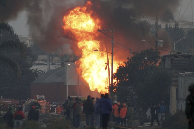 Firefighers and rescue workers stand near a burning gas leak after a series of explosions at a neighborhood in Puebla, Mexico, Sunday, October 31, 2021. Officials say an illegal tap on a gas line is apparently to blame for the early morning explosions that killed at least one person and injured more than a dozen, destroying dozens of homes and causing the evacuation of some 2000 persons. (Photo by Pablo Spencer/AP Photo)