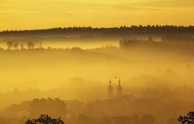 A bird flies past the spires of the church of St. Jude and Simon in Uttenweiler in the morning in the yellowish fog caused by the rising sun in Uttenweiler-Offingen, Germany, Sunday, October 3, 2021. (Photo by Thomas Warnack/dpa via AP Photo)
