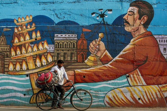 A man carries a bundle of clothes on his bicycle past a wall mural in Varanasi, India on August 19, 2025. (Photo by Niharika Kulkarni/AFP Photo)