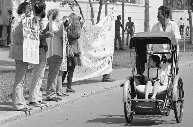 A little girl in a pedicab, and her driver stare as they pass a demonstration of nine anti-war activists before the United States embassy in Saigon, Friday, Jan. 25, 1975. The activists, led by David Harris, left, of Menlo Park, Calif. former husband of folk singer Joan Baez, passed out leaflets demanding the end of U.S. intervention in South Vietnam. (Photo by AP Photo)