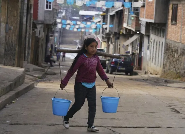 A girl hauling two buckets of water uses a carrying pole in the Puerpecha Indigenous community of Comachuen, Mexico, Wednesday, January 19, 2022.  Money sent home by migrants working in the United States have allowed their families to remain in Comachuen rather than moving to other parts of Mexico for work. That, and the fact kids spend much of the year with their mothers and grandparents, has helped preserve the Purepecha language among almost everyone in town. (Photo by Fernando Llano/AP Photo)
