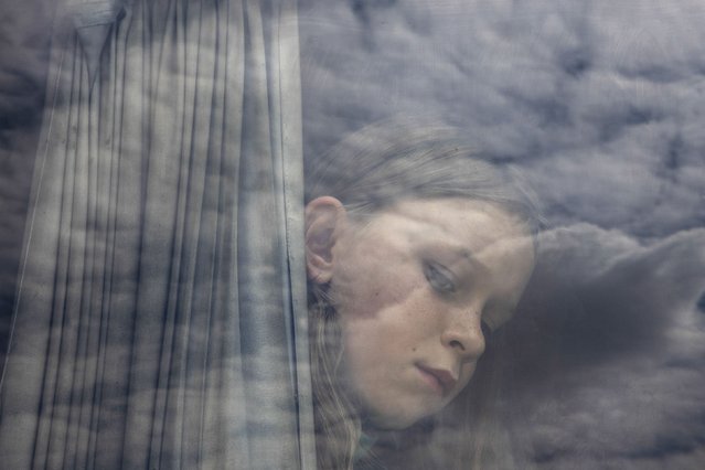 Clouds are reflected in the window of the bus where 10-year-old Polina Budash and her family had gathered for a mandatory evacuation order in Dobropillia, Ukraine, on Saturday, July 26, 2025. Russian forces were approaching the town. (Photo by David Guttenfelder/The New York Times)
