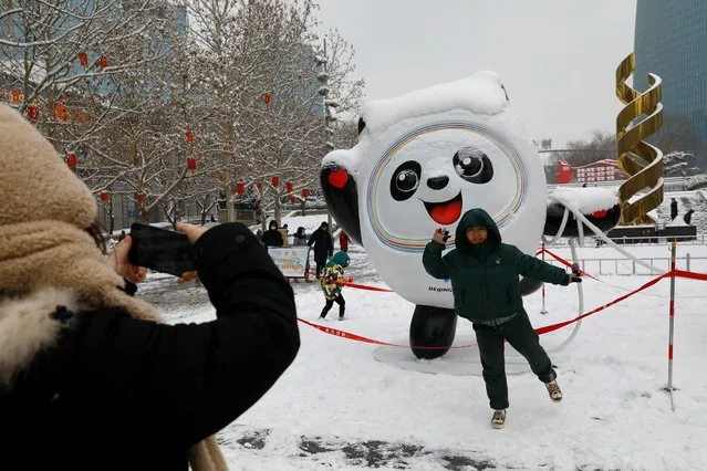 A person poses for pictures in front of an installation featuring Bing Dwen Dwen, the Beijing 2022 Winter Olympic Mascot amidst a snowfall on a street in Beijing, China on February 13, 2022. (Photo by Tingshu Wang/Reuters)