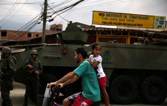 A man rides a bicycle with a girl as they past in front of an armed forces members, during an operation against drug dealers in Vila Kennedy slum in Rio de Janeiro, March 7, 2018. (Photo by Pilar Olivares/Reuters)
