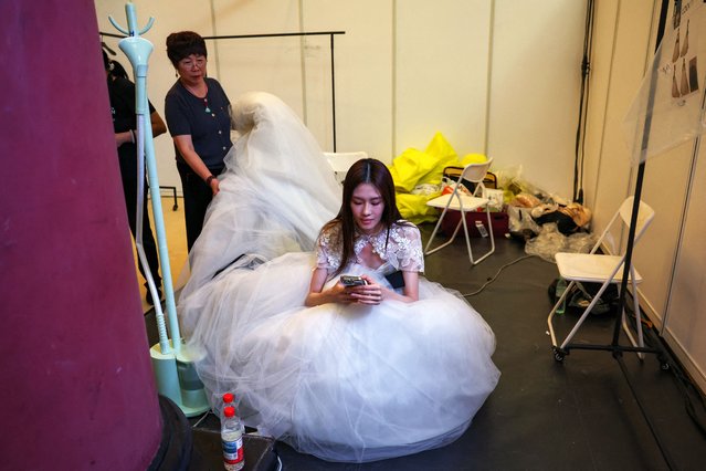 An employee prepares a creation of Vera Wang's Haute Spring/Summer 2026 wedding dress collection as a model waits backstage, at China Fashion Week in Beijing, China on September 10, 2025. (Photo by Tingshu Wang/Reuters)