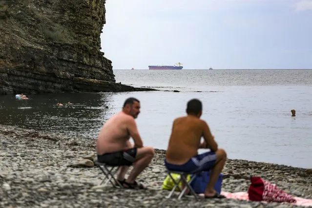Two men rest on the beach as the Minerva Symphony tanker, which sails under the Greek flag is seen at the Black Sea coast after an oil spill, near Novorossiysk, Russia, Thursday, August 12, 2021. Russia'n prosecutors have opened a criminal probe into an oil spill off the country's Black Sea coast that appeared to be far bigger than initially expected. The oil spilled while being pumped into the Minerva Symphony tanker. Authorities initially estimated the spill to cover only 200 square meters (2,153 square feet). But Russian scientists said Wednesday after studying satellite images that it actually covered nearly 80 square kilometers (nearly 31 square miles). (Photo by AP Photo/Stringer)