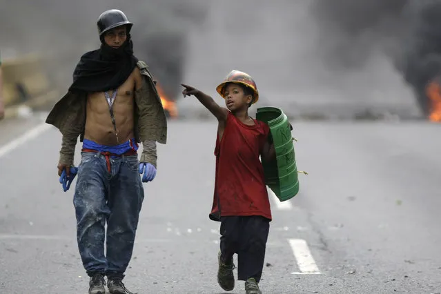 A demonstrator and boy wearing a helmet and holding a shield, walk on a blocked highway as a barricade burns in the background, during a national sit-in against President Nicolas Maduro, in Caracas, Venezuela, Monday, May 15, 2017. Opposition leaders are demanding immediate presidential elections. (Photo by Fernando Llano/AP Photo)
