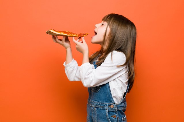 Profile view of a little brunette girl dressed casually and about to bite a slice of pizza in a studio. (Photo by Rex Features/Shutterstock)