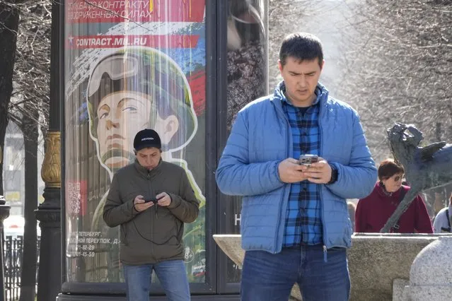 Men look at their phones walking past an army recruiting billboard with the words “Military service under contract in the armed forces”, in St. Petersburg, Russia, Wednesday, April 12, 2023. (Photo by Dmitri Lovetsky/AP Photo)