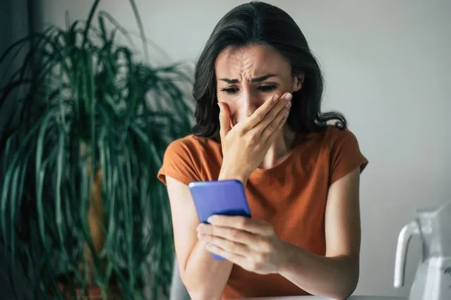 Sad, frustrated young brunette woman is crying with smartphone in hands while she sitting on the chair at apartment. (Photo by Povozniuk/Getty Images)
