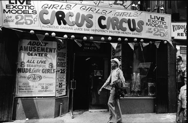 Exterior view of Circus Circus, an 'adult amusement center,' in Times Square as a man walks past wearing a hat and carrying his coat over his arm, New York, New York, 1970s. The marquee advertises exotic models, nude girls, books, magazines, games, and peep shows, as well as a juice bar and 'high-potency protein health drinks