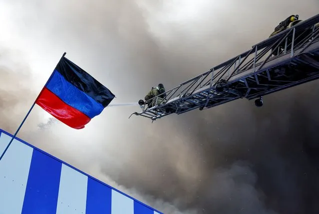 Firefighters work next to a flag of the self-proclaimed Donetsk People's Republic as they attempt to extinguish fire at the shopping mall Galaktika following recent shelling in the course of Ukraine-Russia conflict in Donetsk, Ukraine on August 24, 2022.. (Photo by Alexander Ermochenko/Reuters)