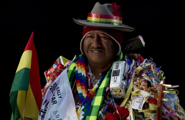 In this February 6, 2018 photo, Lino Reynaldo Chura, dressed as “Ekeko”, the god of prosperity, is covered in objects, including a miniature bus, during a portrait session on the sidelines of the annual Alasita Fair in La Paz, Bolivia. The 64-year-old artisan says his speciality is sewing miniature clothing for Barbies. “I ask the Ekeko for good health, because the rest one attains by working”, he said. Chura competed in this year's Ekeko costume competition. (Photo by Juan Karita/AP Photo)