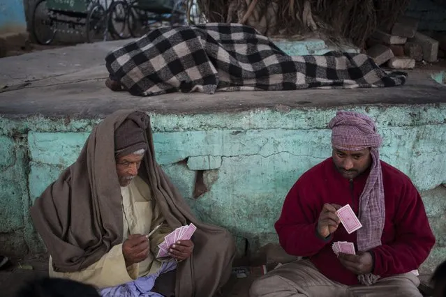 An Indian homeless man sleeps as rickshaw pullers play cards on a cold winter day in New Delhi, India, Wednesday, January 14, 2015. North India continued to reel under cold wave conditions as mercury dipped in several places today with dense fog affecting the movement of traffic, according to local reports. (Photo by Tsering Topgyal/AP Photo)