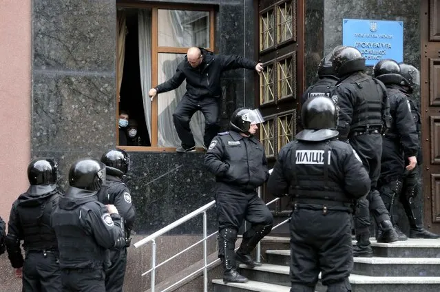 Masked pro-Russian activists look out of a window of the regional prosecutor's office they seized and barricaded inside, as riot police stand, in Donetsk, Ukraine, Saturday, April 12, 2014. Saturday morning a group of pro-Russian activists armed with metal sticks seized the office. They have left the building after talks with police. Nobody was arrested. (Photo by Efrem Lukatsky/AP Photo)
