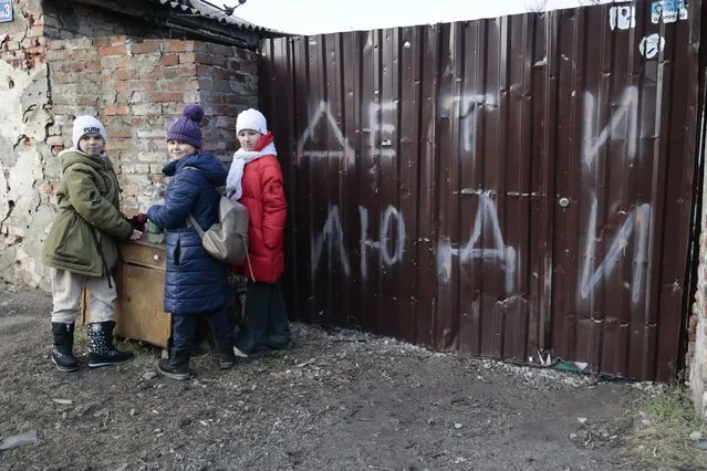 Children stand near the fence of a damaged house with the words "Children and people" after the first anniversary of the Russian Special military operation in Ukraine in Mariupol in Russian-controlled Donetsk region, eastern Ukraine, Saturday, February 25, 2023. (Photo by Alexei Alexandrov/AP Photo)