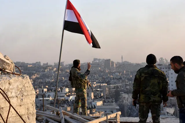 A Syrian government soldier gestures a v-sign under the Syrian national flag near a general view of eastern Aleppo after they took control of al-Sakhour neigbourhood in Aleppo, Syria in this handout picture provided by SANA on November 28, 2016. (Photo by Reuters/SANA)