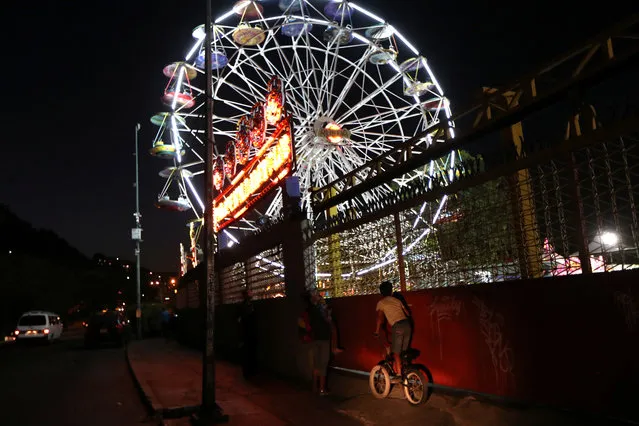 A child climbs on a bike to see an amusement park with an illuminated ferris wheel in Caracas, Venezuela, March 17, 2019. (Photo by Ivan Alvarado/Reuters)