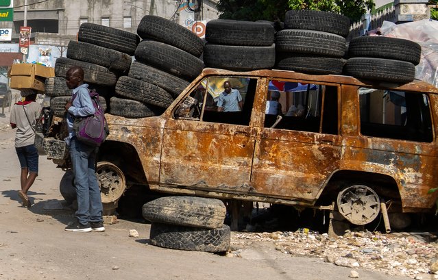 People walk past tires and a burned-out vehicle used to create roadblocks in the Canapé Vert area of Port-au-Prince, Haiti, Monday, April 22, 2024. Haiti's health system has long been fragile, but it's now nearing total collapse after gangs launched coordinated attacks on Feb. 29, targeting critical state infrastructure in the capital and beyond. (Photo by Ramon Espinosa/AP Photo)