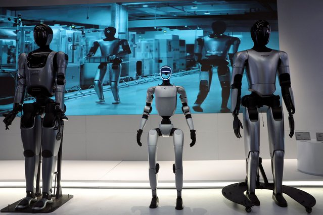Humanoid robots developed by Keenon Robotics, Unitree Robotics and MagicLab stand in front a display showing video of humanoid robots' application in factories, during an organised media tour to Robot Mall, a store selling humanoid and other robots, in Beijing, China on August 6, 2025. (Photo by Florence Lo/Reuters)