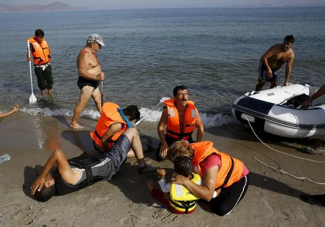 A tourist walks past an Iranian migrant who collapsed next to his son and wife moments after a small group arrived by engineless dinghy from the Turkish coast (seen in the background) at a beach on the Greek island of Kos August 15, 2015. (Photo by Yannis Behrakis/Reuters)