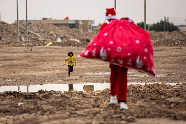 A child runs towards Mohamed Maarouf, 28, as he walks with a sack of gifts while dressed in Saint Nicholas (Santa Claus) costume in a slum near the centre of Iraq's southern city of Basra on December 24, 2021. (Photo by Hussein Faleh/AFP Photo)
