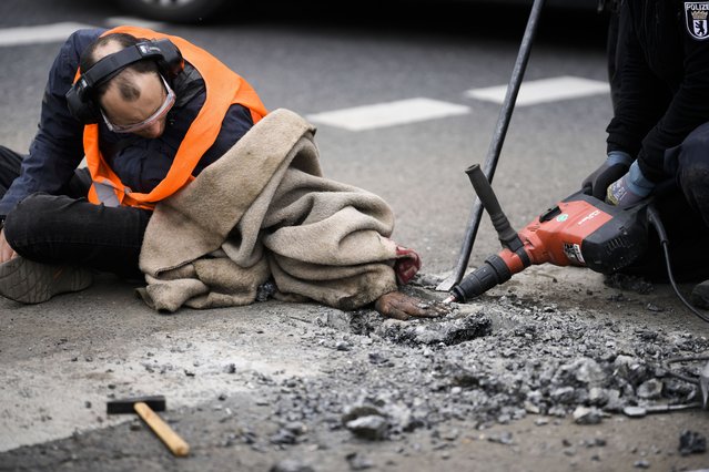 Police remove the asphalt next to the hand of an climate activist who glued his hand on the road, during a protest against the climate policy of the German government in Berlin, Germany, Monday, April 24, 2023. German climate activists tried bringing traffic to a standstill in Berlin on Monday morning by gluing themselves to streets all over the capital. (Photo by Markus Schreiber/AP Photo)