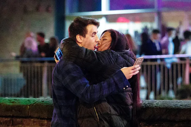A couple embrace and kiss while using their mobile phones during a pre-Christmas “Mad Friday” night in Manchester, England. This unposed scene is reminiscent of Banksy’s “Mobile Lovers”. (Photo by Joel Goodman/The Guardian)