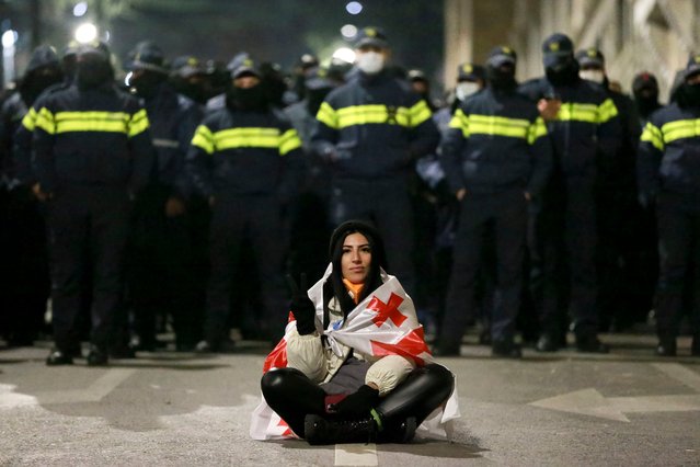 A demonstrator draped in a Georgian national flag sits in front of police rallying outside the parliament's building to continue protests against the government's decision to suspend negotiations on joining the European Union in Tbilisi, Georgia, on Monday, December 2, 2024. (Photo by Zurab Tsertsvadze/AP Photo)