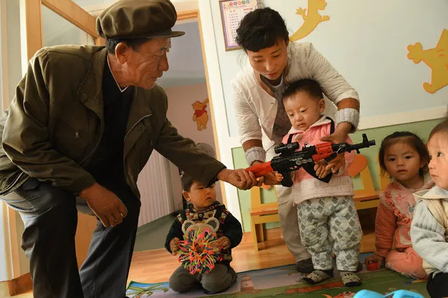 A 2- 3 year-old-child holds a toy machine gun during a media tour of a day care / preschool center at Jangchon cooperative farm on the outskirts of  Pyongyang, North Korea on May 4, 2016. Man at left is said to be a farmer there. The homes had vegetable gardens in the front, solar panels on the roofs but didn't appear to be very populated. (Photo by Linda Davidson/The Washington Post)