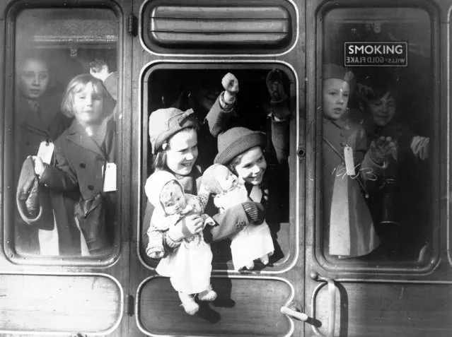 Two small girls hold their dolls while waving goodbye to relatives from a train carriage as they are evacuated to the west country, at a London station, to escape the Nazi bombing, June 13, 1940. All the children wear identity labels. (Photo by AP Photo)