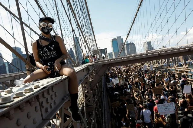 A woman wearing a face mask sits on a metal fence as people march during events to mark Juneteenth, which commemorates the end of slavery in Texas, two years after the 1863 Emancipation Proclamation freed slaves elsewhere in the United States, amid nationwide protests against racial inequality, at the Brooklyn Bridge, in New York City, New York, U.S., June 19, 2020. (Photo by Brendan Mcdermid/Reuters)