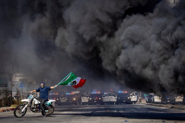 A man on a motorcycle waves a Mexican flag as smoke rises from a burning car on Atlantic Boulevard, during a standoff by protesters and law enforcement, following multiple detentions by Immigration and Customs Enforcement (ICE), in the Los Angeles County city of Paramount, California, U.S., June 7, 2025. (Photo by Barbara Davidson/Reuters)