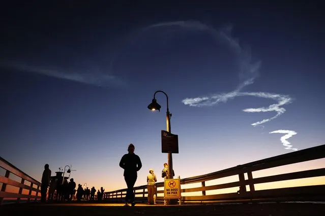 People walk on a pier beneath the contrail remaining from a SpaceX Falcon 9 rocket carrying a payload of 22 Starlink internet satellites into space after launching from Vandenberg Space Force Base on April 1, 2024 in San Clemente, California. The launch was visible across much of Southern California. (Photo by Mario Tama/Getty Images)