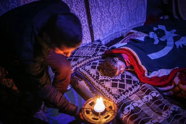 A Syrian father is seen near his child sleeping at a makeshift tent during cold weather in Idlib, Syria on January 24, 2022. Fathers in Syria's northwestern province of Idlib wait all night in the freezing cold so that their children do not freeze. Civilians living in tents in the city, where thermometers show sub-zero at night, have difficulty in warming. (Photo by Muhammed Said/Anadolu Agency via Getty Images)