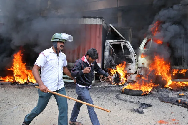 An Indian police officer detains a youth as they walk past burning trucks that belong to the neighboring Tamil Nadu state set ablaze by angry mobs in Bangalore, Karnataka state, India, Monday, September 12, 2016. India's top court on Monday ordered the southern state of Karnataka to release water from a disputed river to neighboring Tamil Nadu after violence erupted in both states over water sharing. (Photo by Aijaz Rahi/AP Photo)