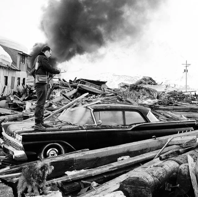 In this March 29, 1964 file photo, a photographer looks over wreckage as smoke rises in the background from burning oil storage tanks at Valdez, Alaska. On Monday, February 1, 2016, federal scientists say they've pinpointed the cause of tsunami waves following the 1964 Great Alaska Earthquake, the second-largest ever recorded, at magnitude 9.2. (Photo by AP Photo)