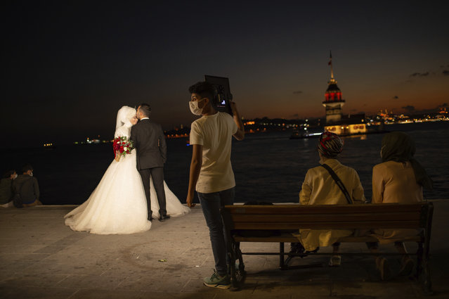A wedding photographer's assistant, wearing a mask for protection against the spread of coronavirus, holds lighting equipment, as a couple poses for photographs backdropped by Istanbul's iconic Maiden's Tower (Kiz Kulesi) at the Bosphorus Strait separating the European and Asian sides of Istanbul, Friday, September 11, 2020. (Photo by Yasin Akgul/AP Photo)